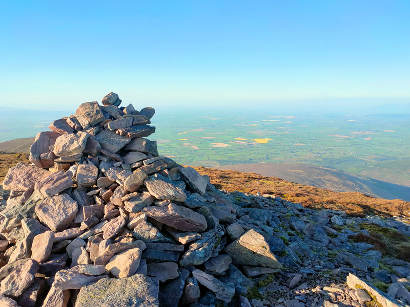 Cairn of stones on top of Knockmoylan, patchwork of Green and yellow fields reaching out to white/blue sky