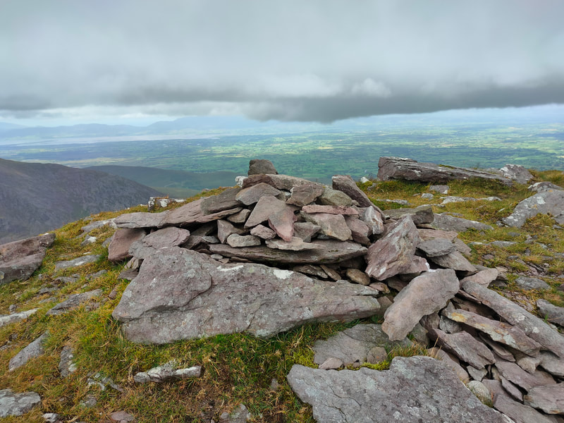 A few stones marking the top of Maolán Buí with views of county Kerry with dark low hanging clouds