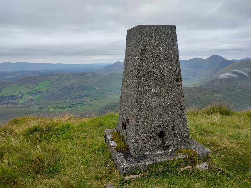 Mullaghanattin Trig Pillar on a grassy mountain top with more mountains in the background