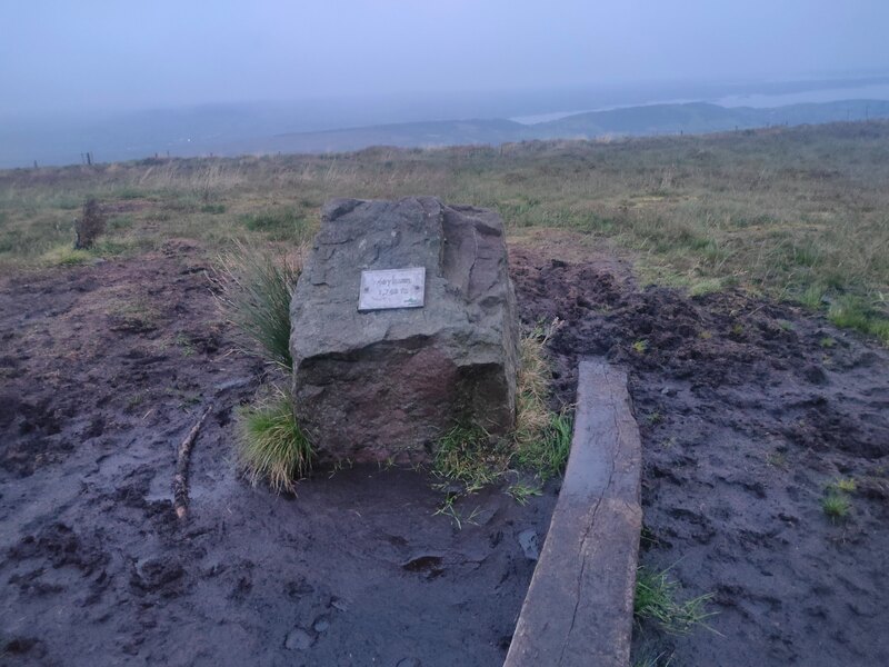 A large stone and plaque mark the top of Moylussa SE, surrounded by mud and a plank for access.