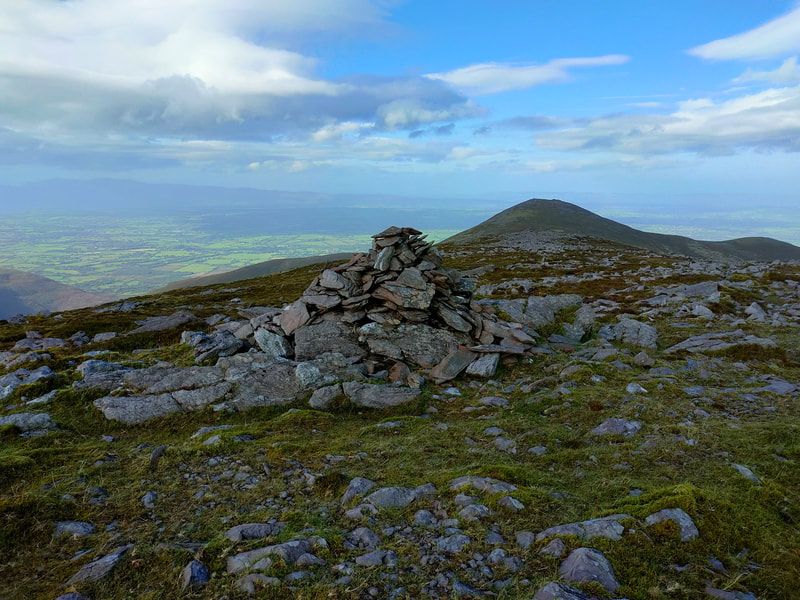 A small pile of stones at the top of Purple Mountain Northeast with Tomies Mountain beyond against an bright blue cloudy sky.