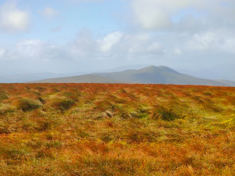A brown grassy mountain top looking across at a twin peaked mountain against a blue sky with some white clouds