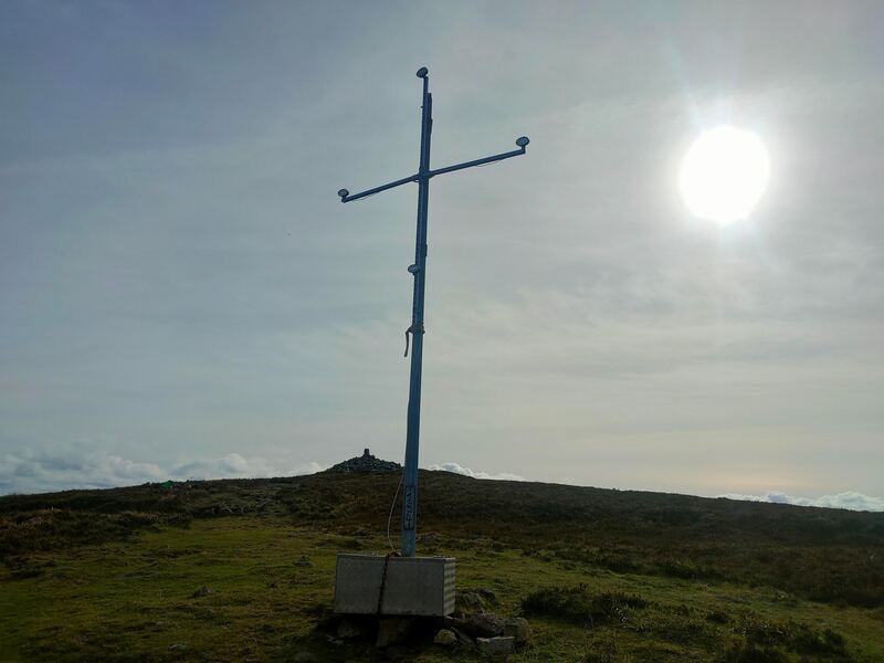 A steel cross on Brandon Hill, white sun trying to get through the clouds. Trig Pillar and cairn of stones behind the cross