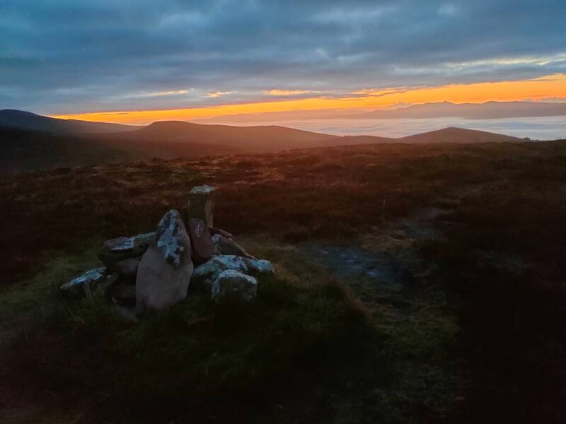 A few stones on the top of Monabrack, oranges and blues of a sunrise sky in the background with a cloud inversion in the valley
