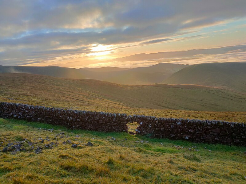 Dry stone wall with hole on grassy mountainside. Early morning sun shine and shadows on distant mountain ranges.