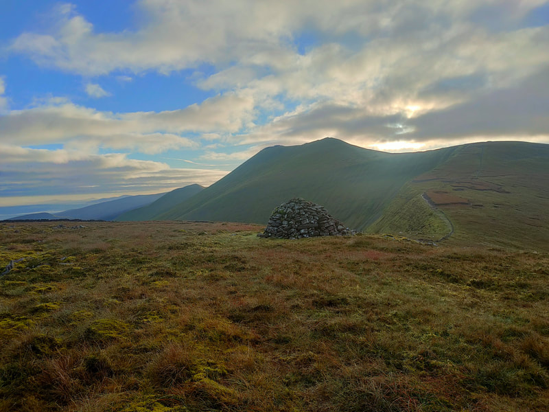 Mound of stones on grassy mountain top, path leading up the mountain edge